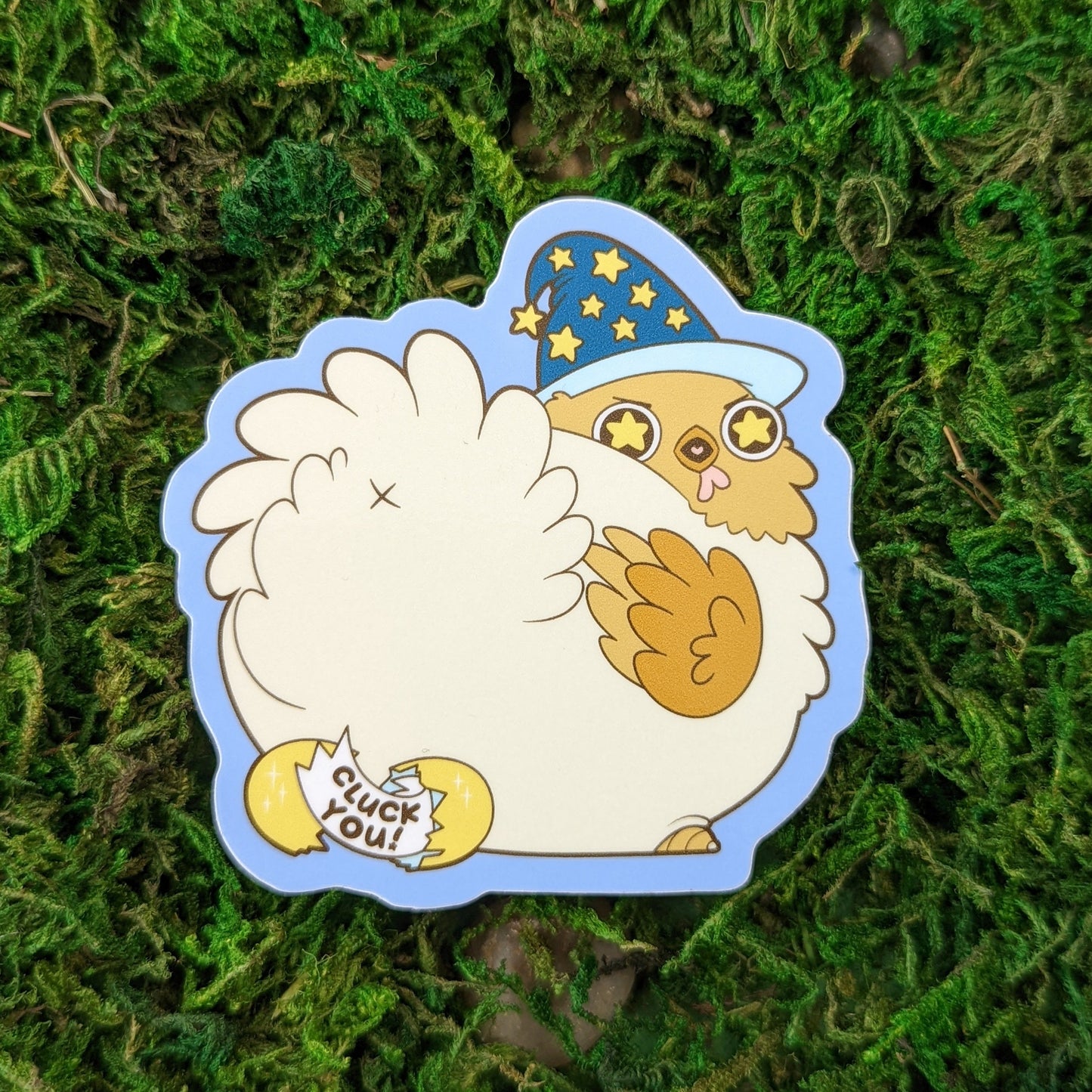 Photograph of a mossy background with a vinyl sticker of a chicken with a wizard hat. A hatched gold egg behind it has a slip of paper in it that says, “Cluck You!”