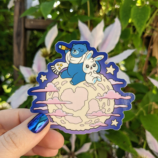 A photograph of a vinyl sticker being held in front of a clematis plant. The sticker is a pink moon with Ursa the blue star bear and Mochi the white moon bunny star gazing on top of the moon.