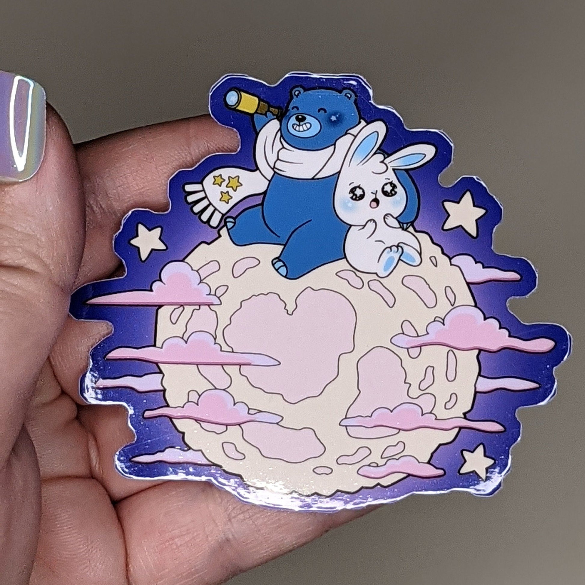 A photograph of a vinyl sticker being held.The sticker is a pink moon with Ursa the blue star bear and Mochi the white moon bunny star gazing on top of the moon.