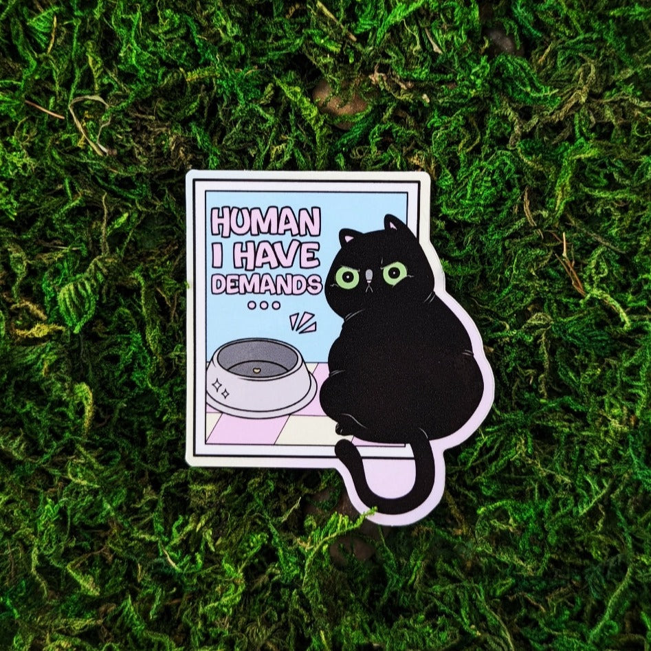 Photograph of a mossy background with a vinyl sticker of a black cat looking backwards sternly. Next to the cat on a pink and yellow tiled floor is a cat food bowl with a single piece of cat food. Above the bowl it says, “Human, I have demands.”