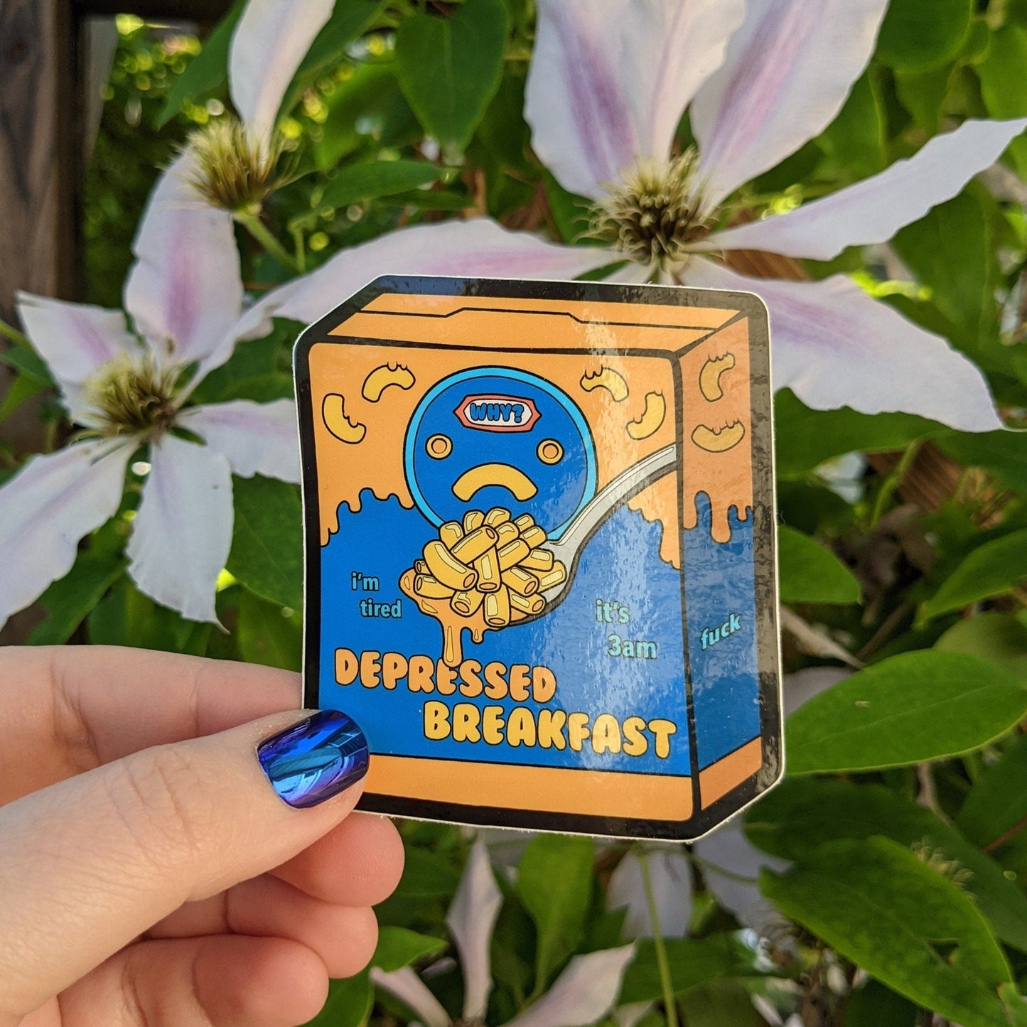 A photograph of a vinyl sticker being held in front of a clematis plant. The vinyl sticker is a box of mac and cheese that says: “Depressed Breakfast.”