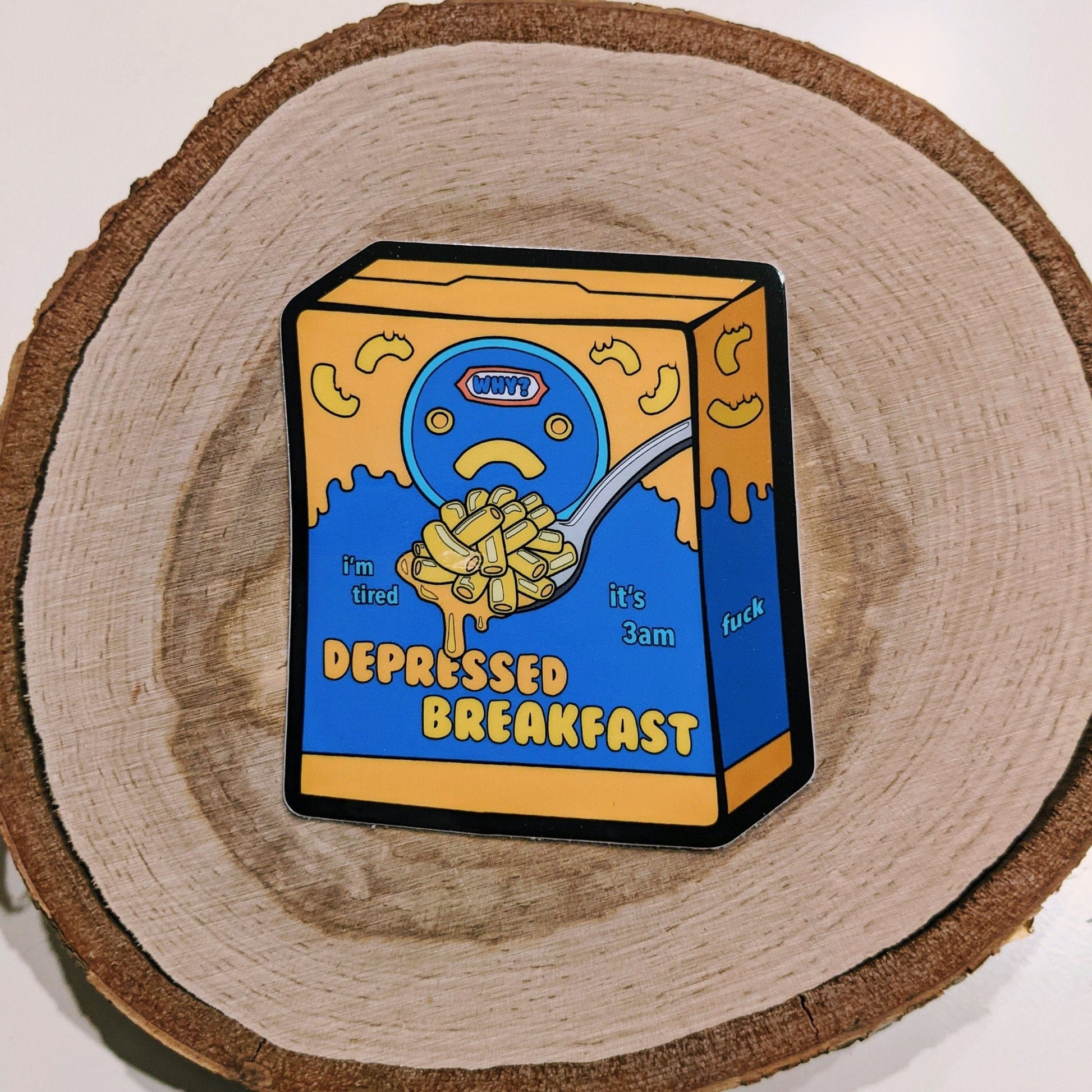 A photograph of a white background and a sticker on a birch round. The vinyl sticker is a box of mac and cheese that says: “Depressed Breakfast.”