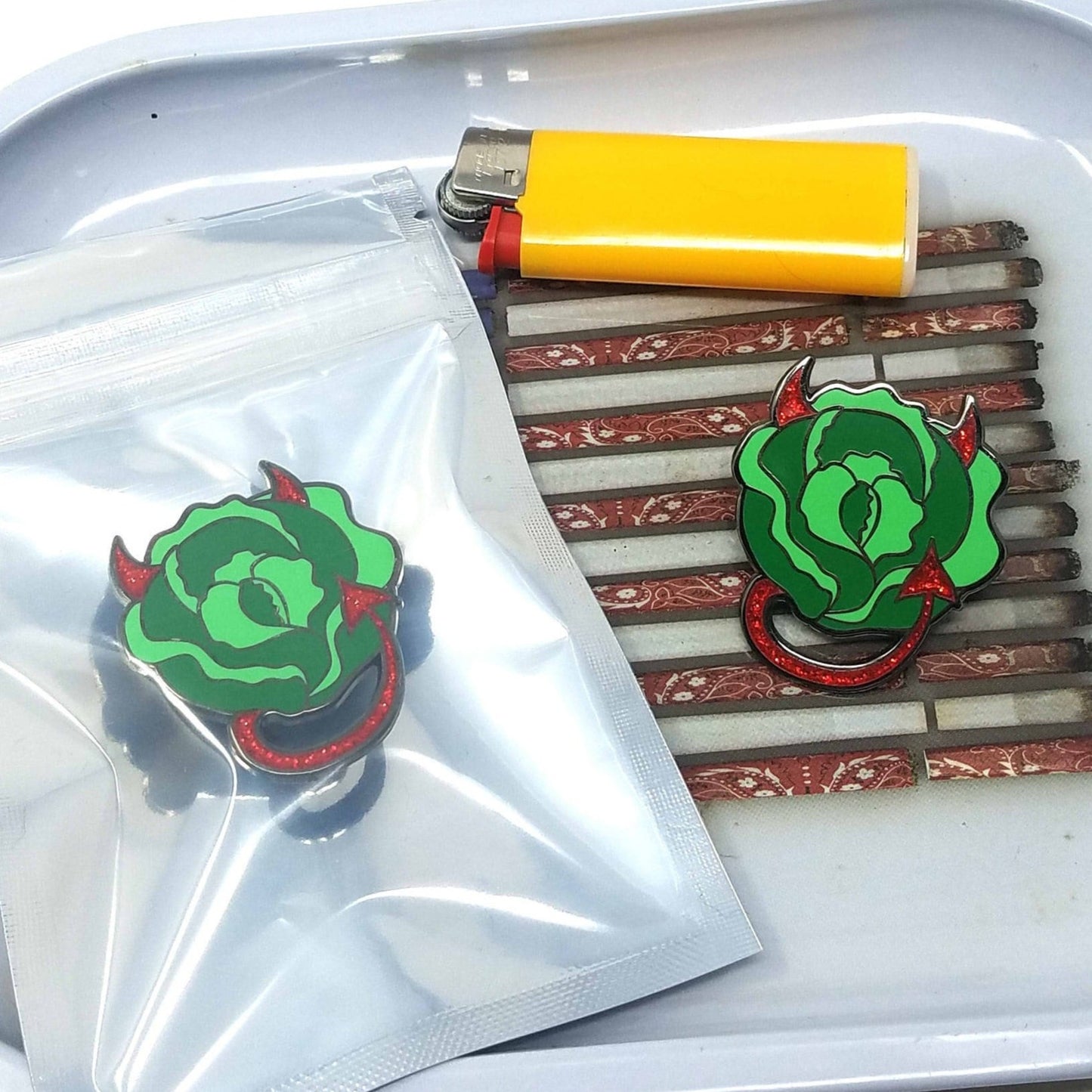 A photograph of a rolling tray with a lighter and two enamel pins. The pins are lettuce with glittery devil horns and tail.