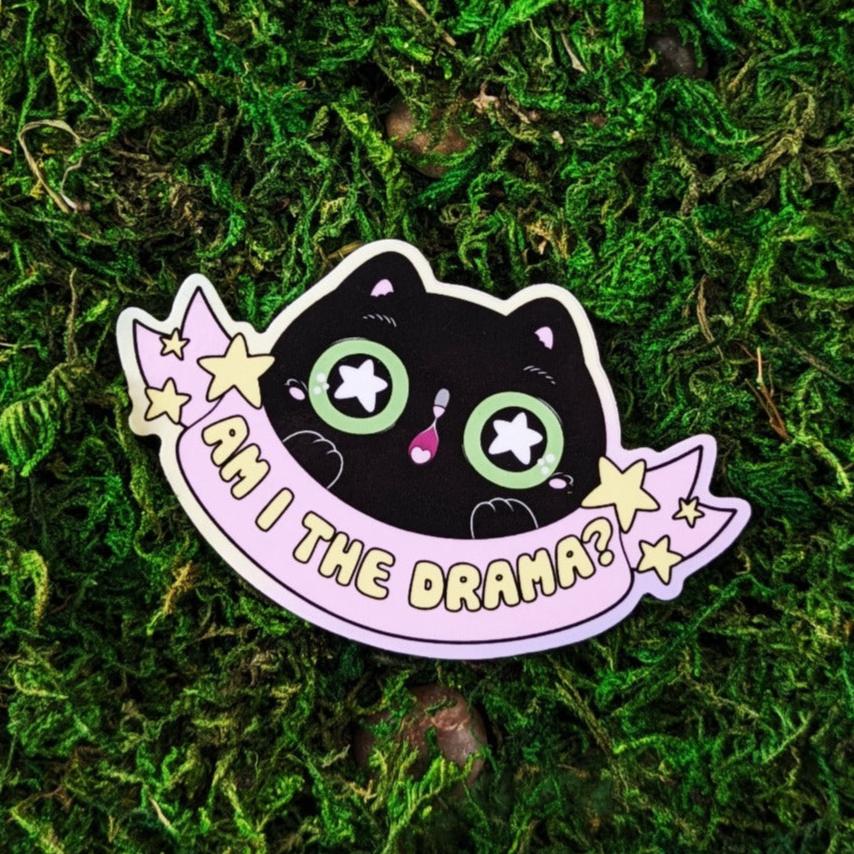 Photograph of a mossy background with a vinyl sticker of a black cat with green eyes. The cat appears to be gasping in surprise. Under the cat is a pink banner that says, “Am I the Drama?”