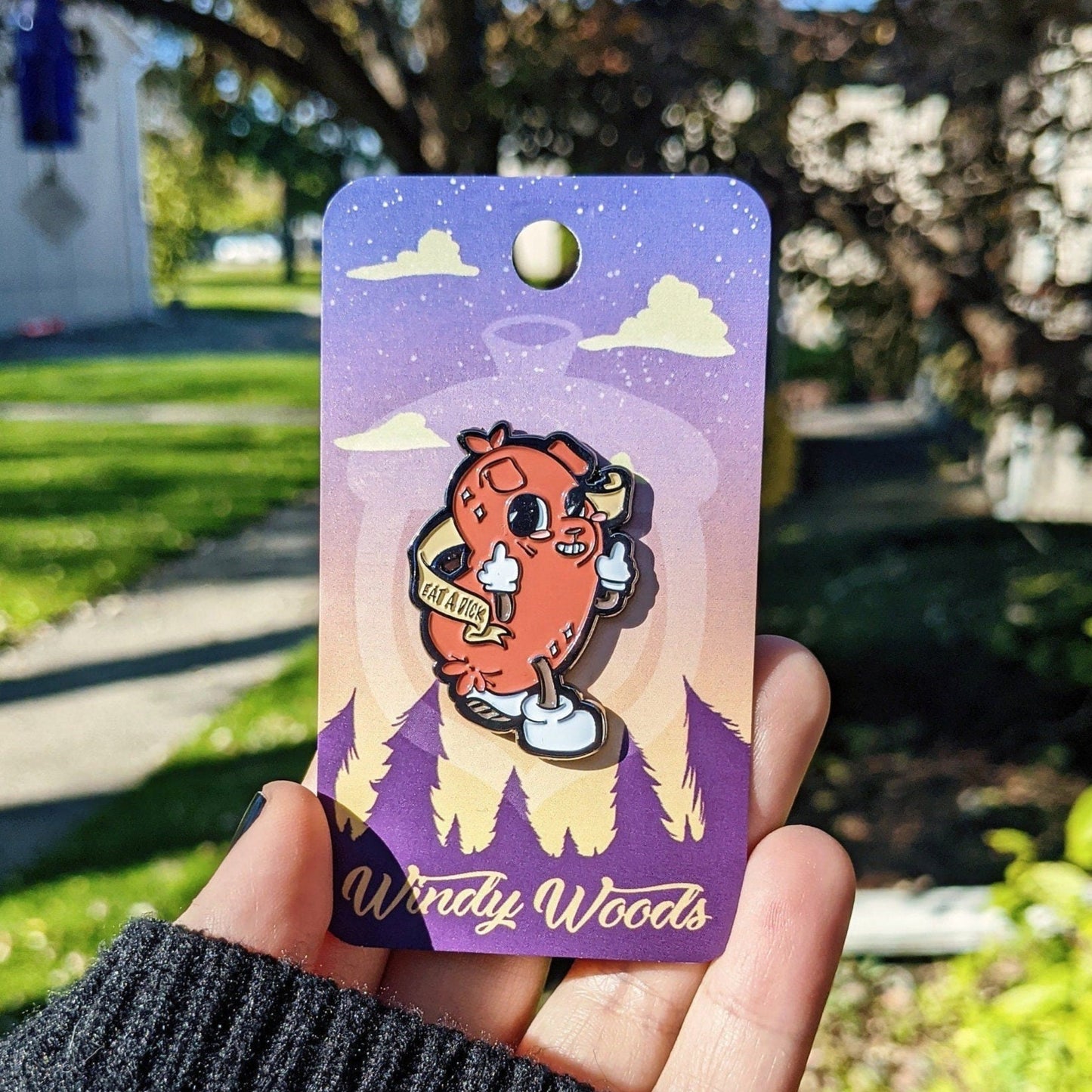 A photograph of someone holding an enamel pin on a backing card that says Windy Woods. The enamel pin is of a cartoon dog/ hot dog hybrid while flipping the bird. Around the dog is a scroll banner that says "Eat a dick."