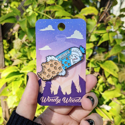 A photograph of an enamel pin being held in front of a clematis plant. The enamel pin is of a roll of cookie dough that says forbidden candybar.