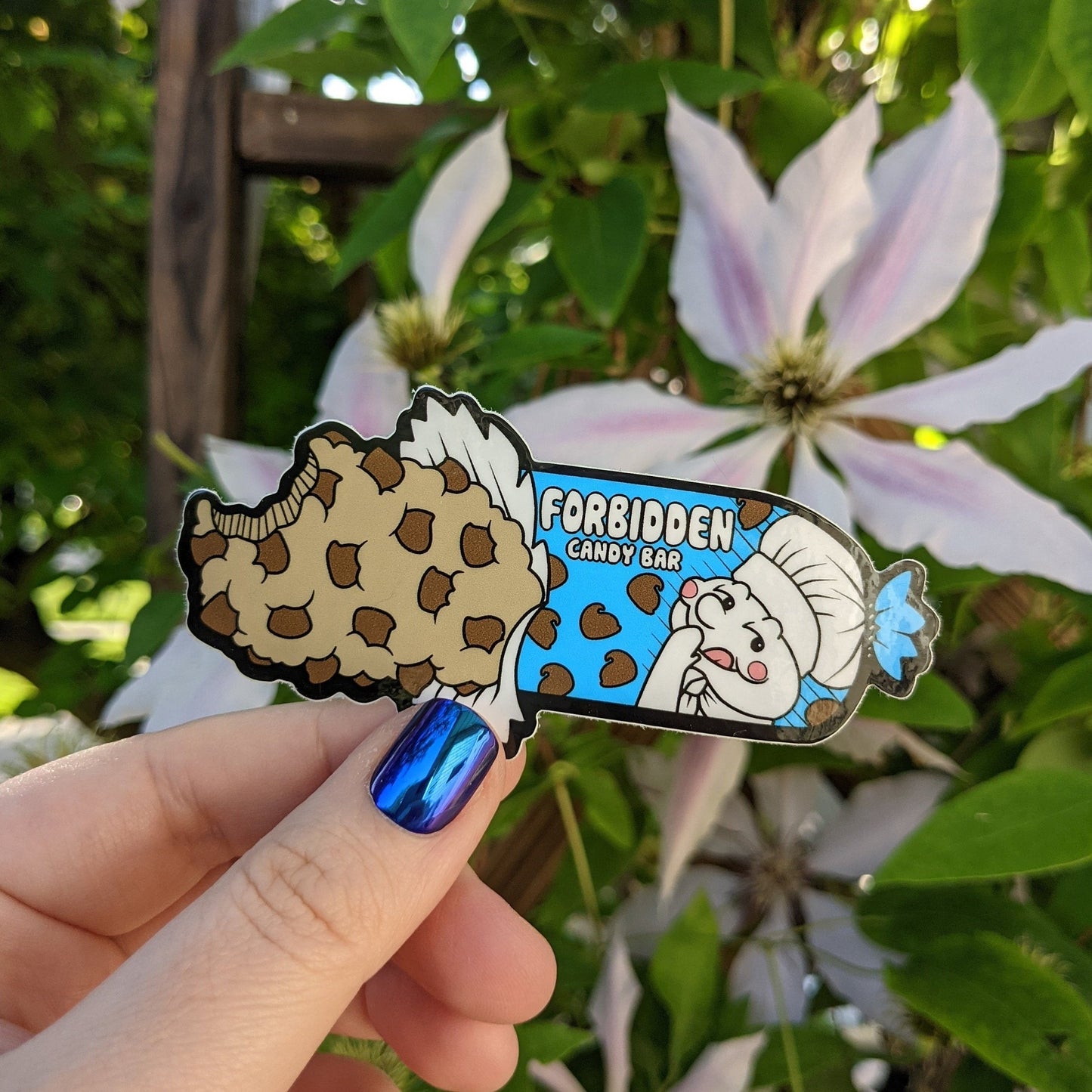 A photograph of a vinyl sticker being held in front of a clematis plant. The vinyl sticker is a package of cookie dough that says forbidden candy bar.