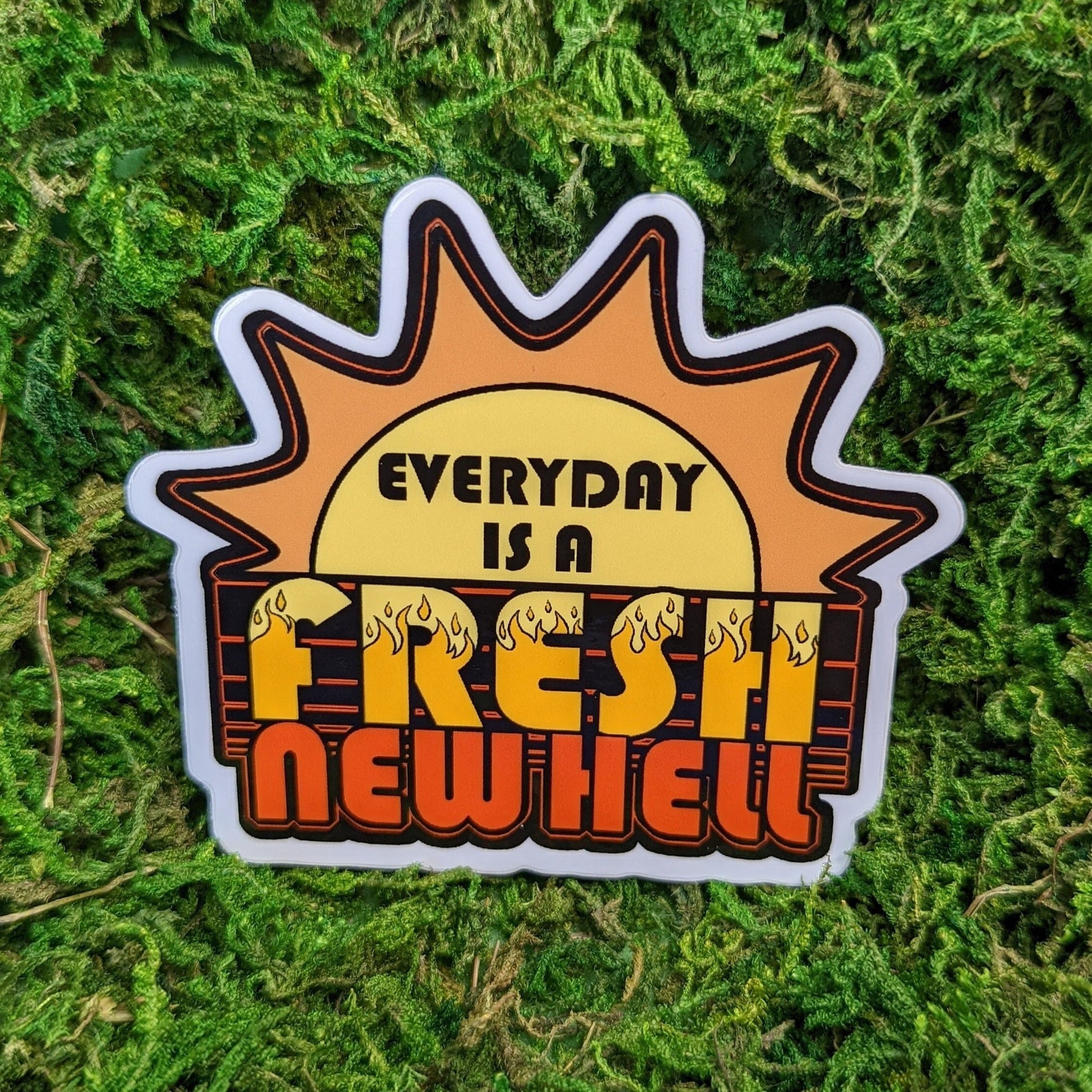 Photograph of a mossy background with a vinyl sticker of a graphic sun in an 80’s style. In front of the sun it says, “Everyday is a fresh new hell.”