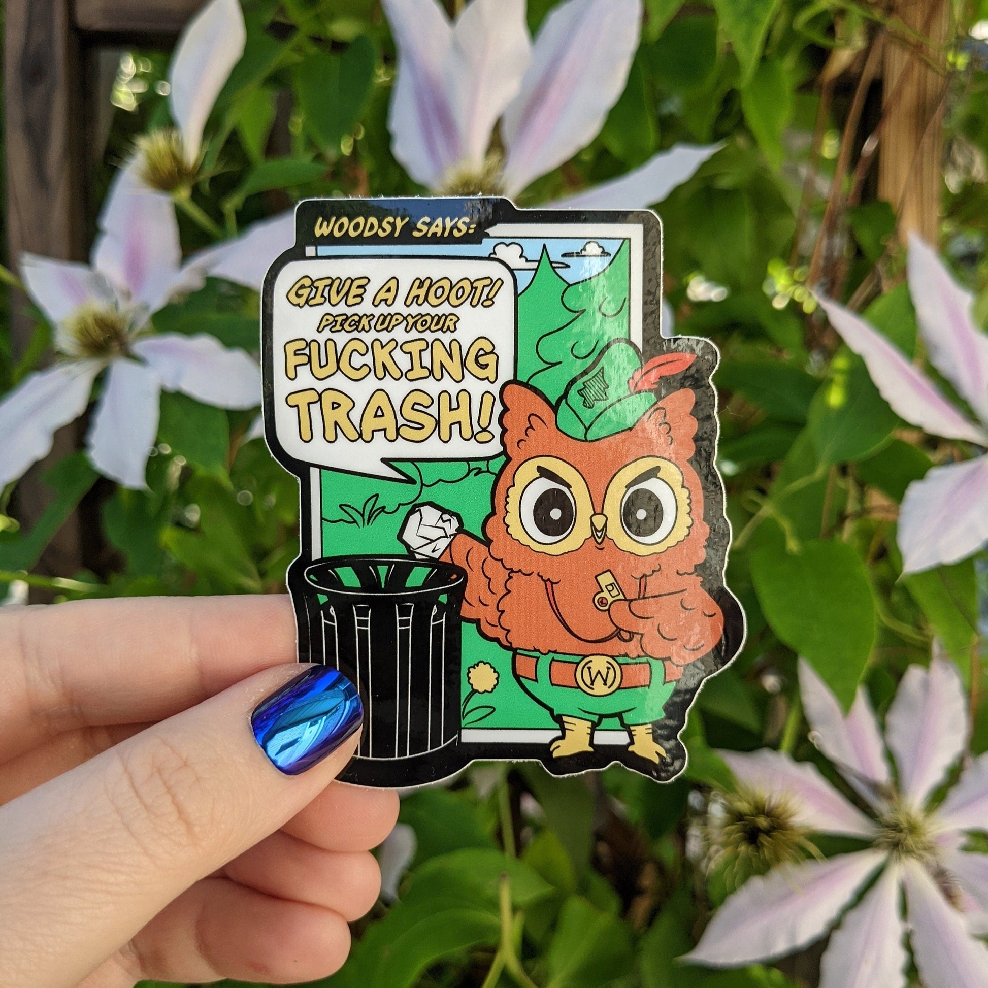 A photograph of a vinyl sticker being held in front of a clematis plant. The sticker is of Woodsy the owl with a whistle putting a piece of trash into a garbage can. Woodsy is saying: “Give a hoot! Pick up your fucking trash!”