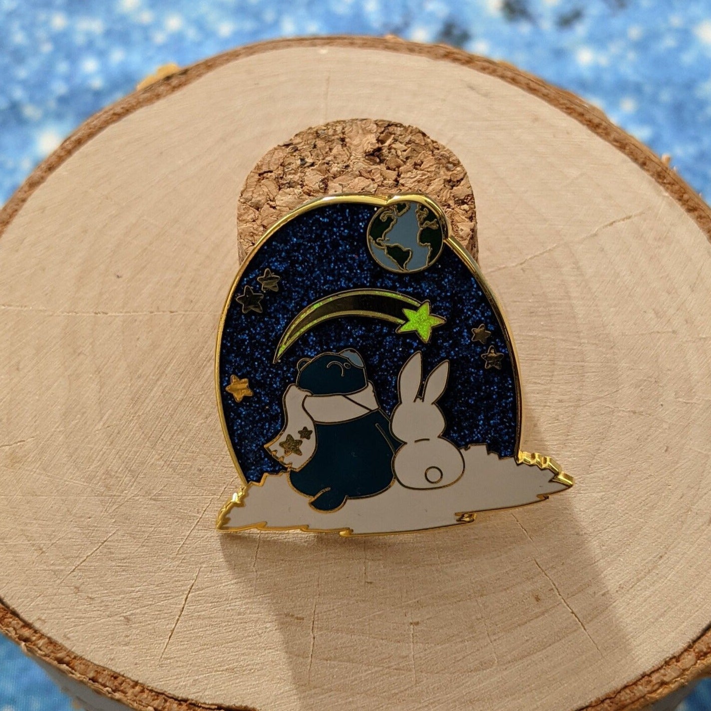 A photograph of a birch round and a cork holding up an enamel pin. The enamel pin artwork is of a blue star bear in a scarf and a white rabbit looking up at the Earth and a shooting star from the surface of the moon.