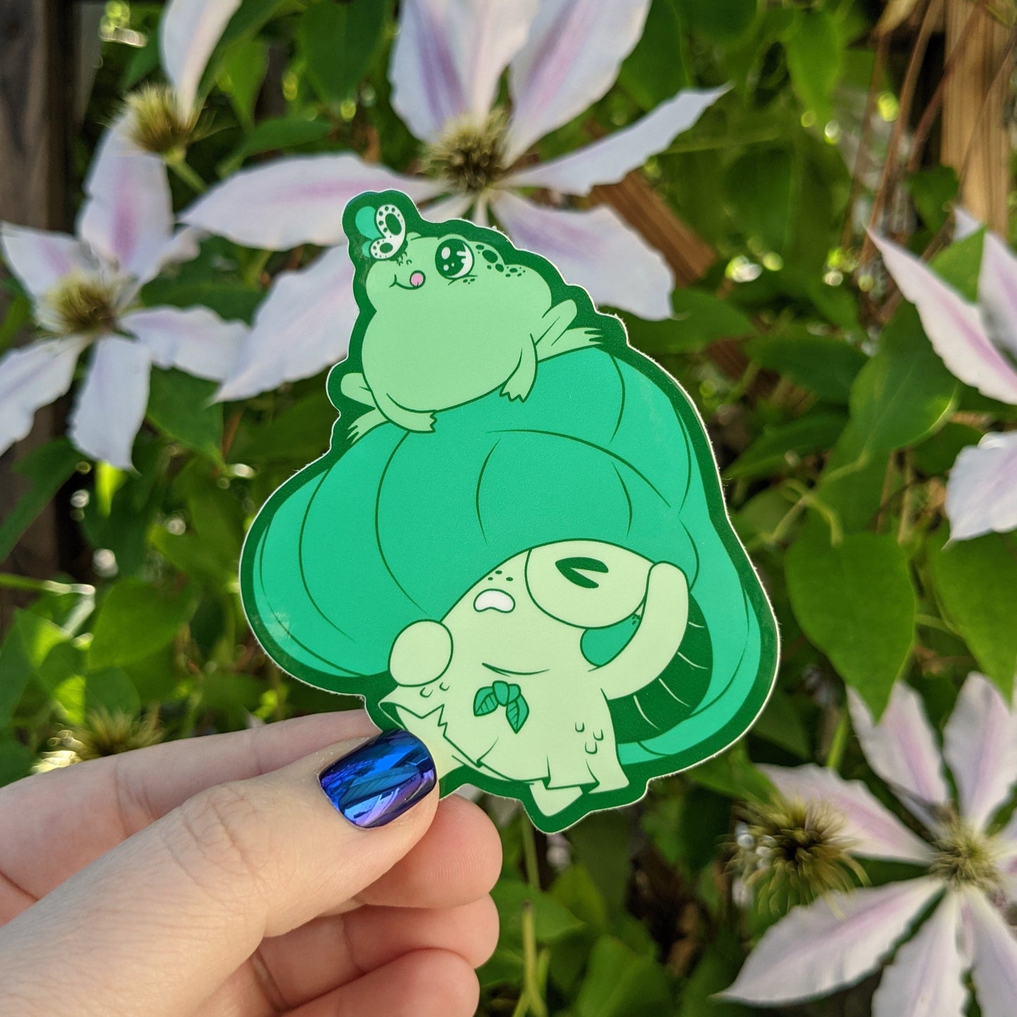 A photograph of a vinyl sticker being held in front of a clematis plant. The sticker is of a green mushroom sprite called a Mushkin. Mushkin is scared because a green frog has jumped on their cap.