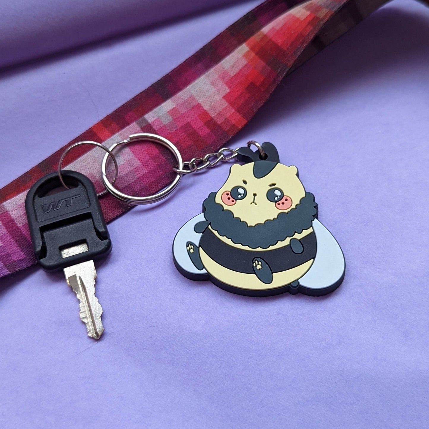 A photograph of a purple background with a landyard and a key. Attached to the key is a PVC Keychain of a hamster bumble bee hybrid creature called a Honeyham.