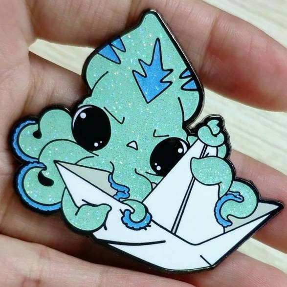 an enamel pin of a light blue squid grabbing onto a paper boat.