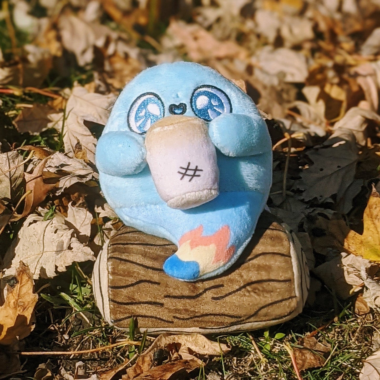 A photograph of Mallow the Campfire Ghost Plush. Mallow is sitting on some foliage. Mallow has a log he is sitting on, a little flame at the tip of his ghost tail, and he's holding a toasty marshmallow. 