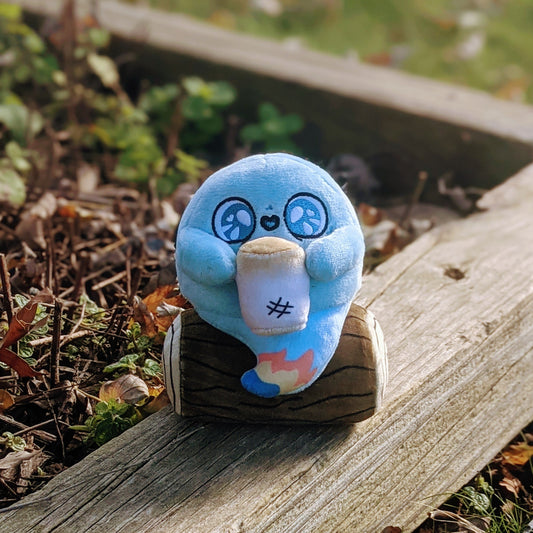 A photograph of Mallow the Campfire Ghost Plush. Mallow is sitting on a wood beam by some foliage. Mallow has a log he is sitting on, a little flame at the tip of his ghost tail, and he's holding a toasty marshmallow. 