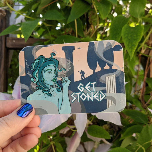 A photograph of a vinyl sticker being held in front of a clematis plant. The vinyl sticker is of Medusa smoking a joint with silhouettes of pillars and men turned to stone in the background. Next to Medusa it says, “Get stoned.”