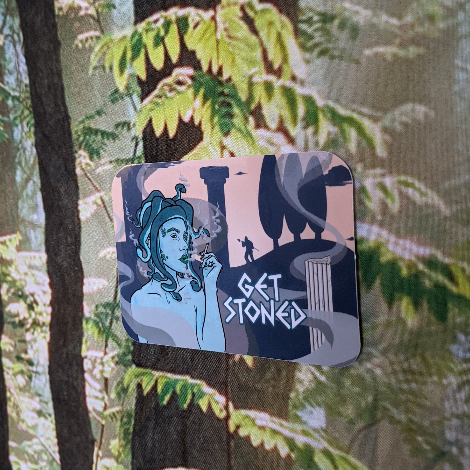 A photograph of a vinyl sticker being held in front of a clematis plant. The vinyl sticker is of Medusa smoking a joint with silhouettes of pillars and men turned to stone in the background. Next to Medusa it says, “Get stoned.”