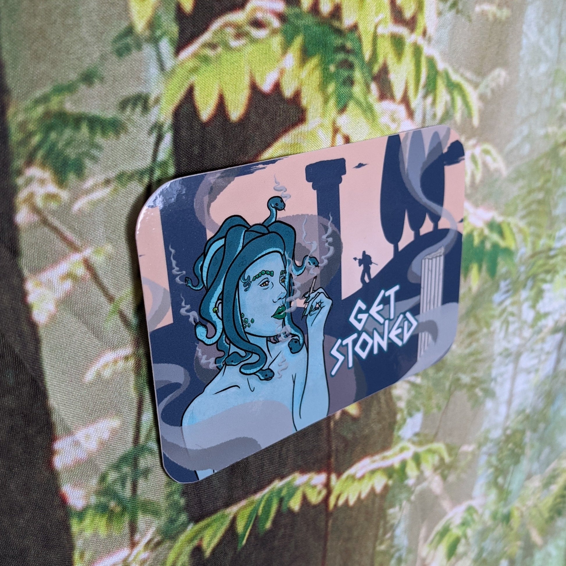 A photograph of a vinyl sticker being held in front of a clematis plant. The vinyl sticker is of Medusa smoking a joint with silhouettes of pillars and men turned to stone in the background. Next to Medusa it says, “Get stoned.”