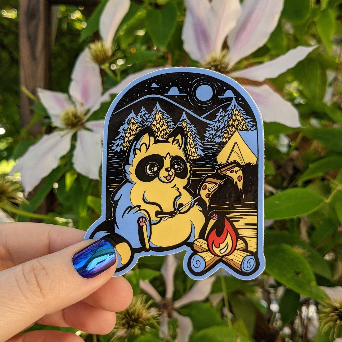 A photograph of a vinyl sticker being held in front of a clematis plant. The vinyl sticker is a raccoon roasting pizza over a campfire. The scene is colored in all yellows	and blues. Behind the raccoon is a night forest and a full moon.