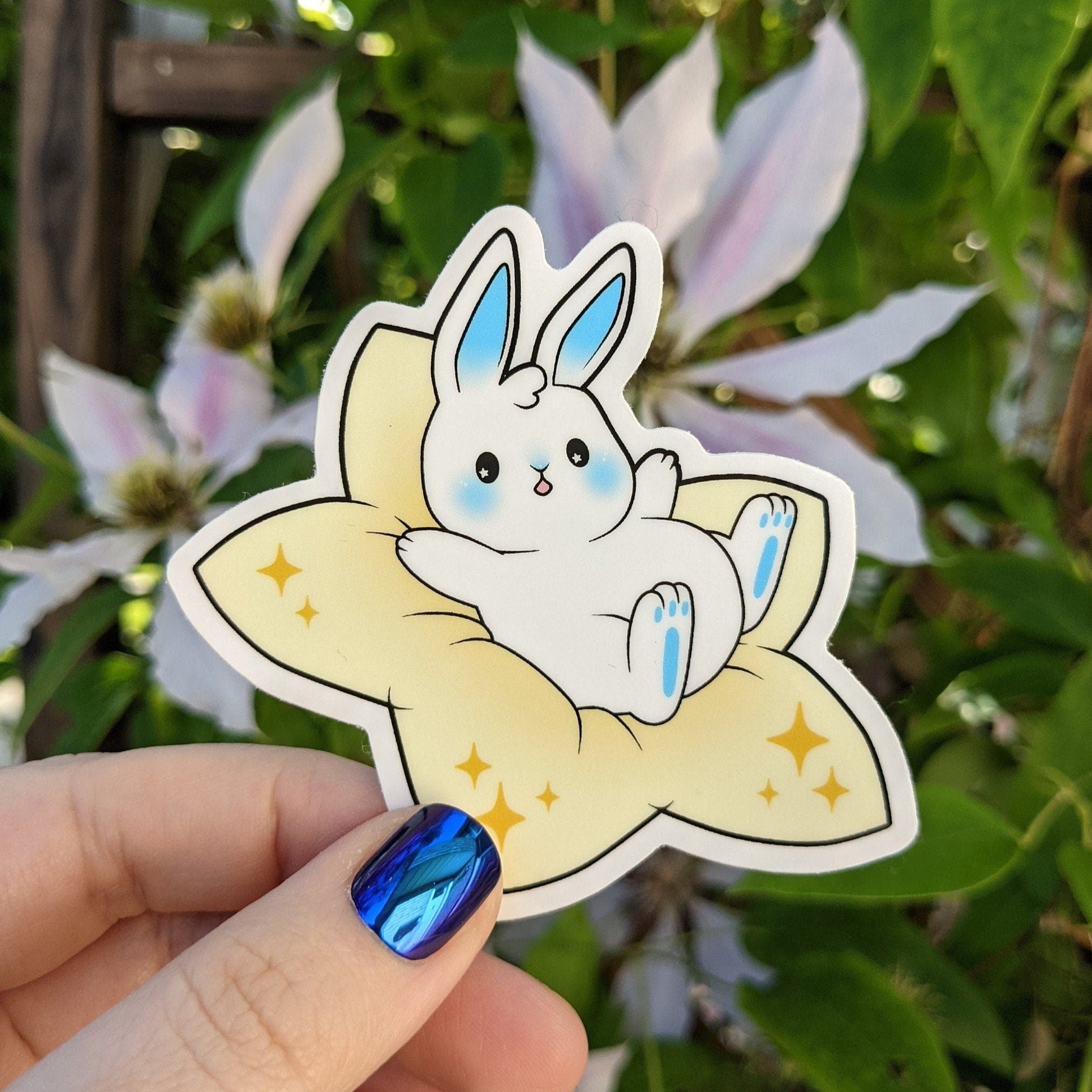 A photograph of a vinyl sticker being held in front of a clematis plant. The vinyl sticker of a white bunny with blue accents. The bunny is sitting on a plush star.