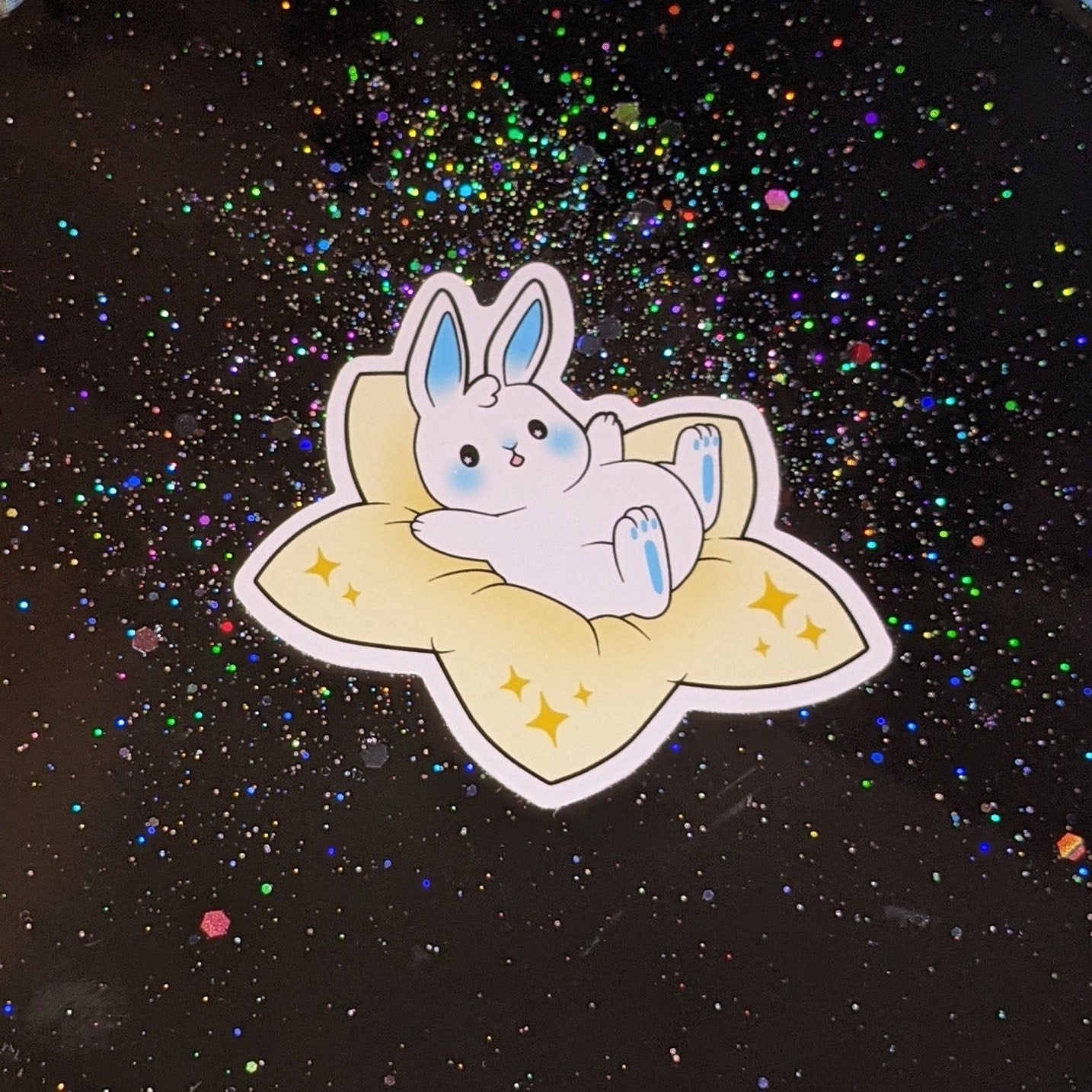 A photograph of a black background with glitter sparkles. On the background is a photo of a vinyl sticker of a white bunny with blue accents. The bunny is sitting on a plush star.