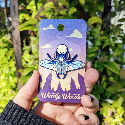 Photograph of an enamel pin being held in front of some plants. The enamel pin is of a beetle with moon themes.