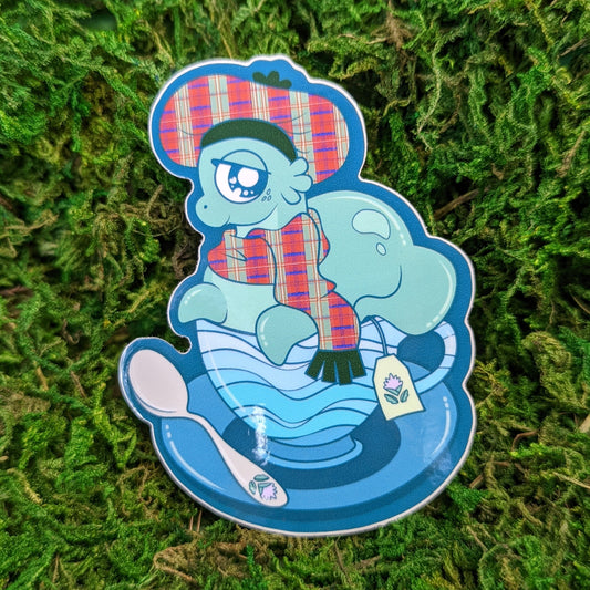 Photograph of a mossy background with a vinyl sticker of the cryptid Nessie. Nessie is sitting in a water themed tea cup while wearing a tartan beret and scarf.
