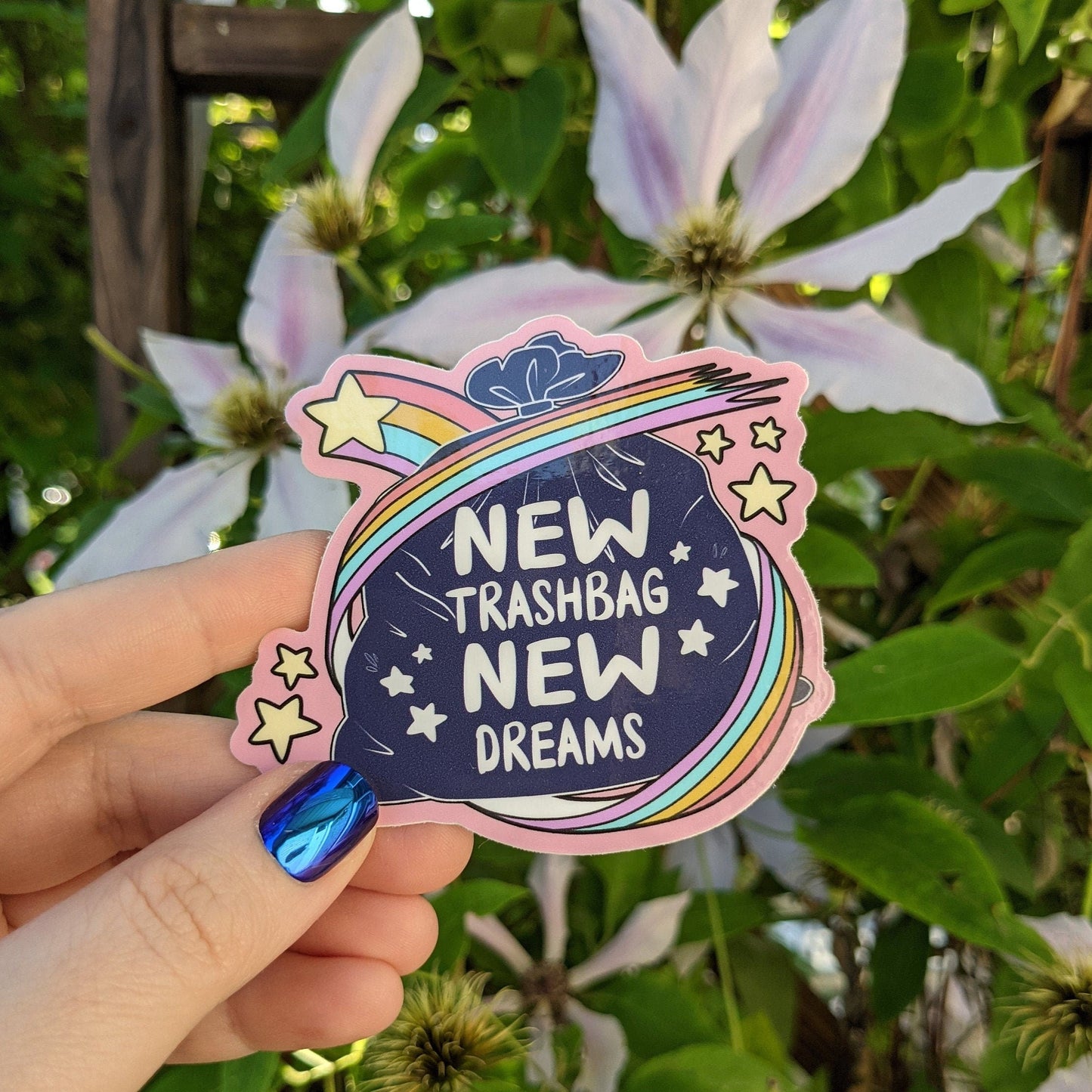 Photograph of a mossy background with a vinyl sticker of a trash bag with rainbow stars around it. The trash bag says, “New Trash Bag New Dreams.”