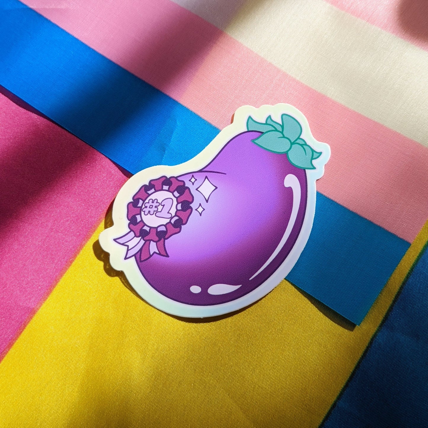 A photograph of a background with the transgender and pansexual pride flags. On top of those is a vinyl sticker of an eggplant with a sparkly #1 ribbon on it.