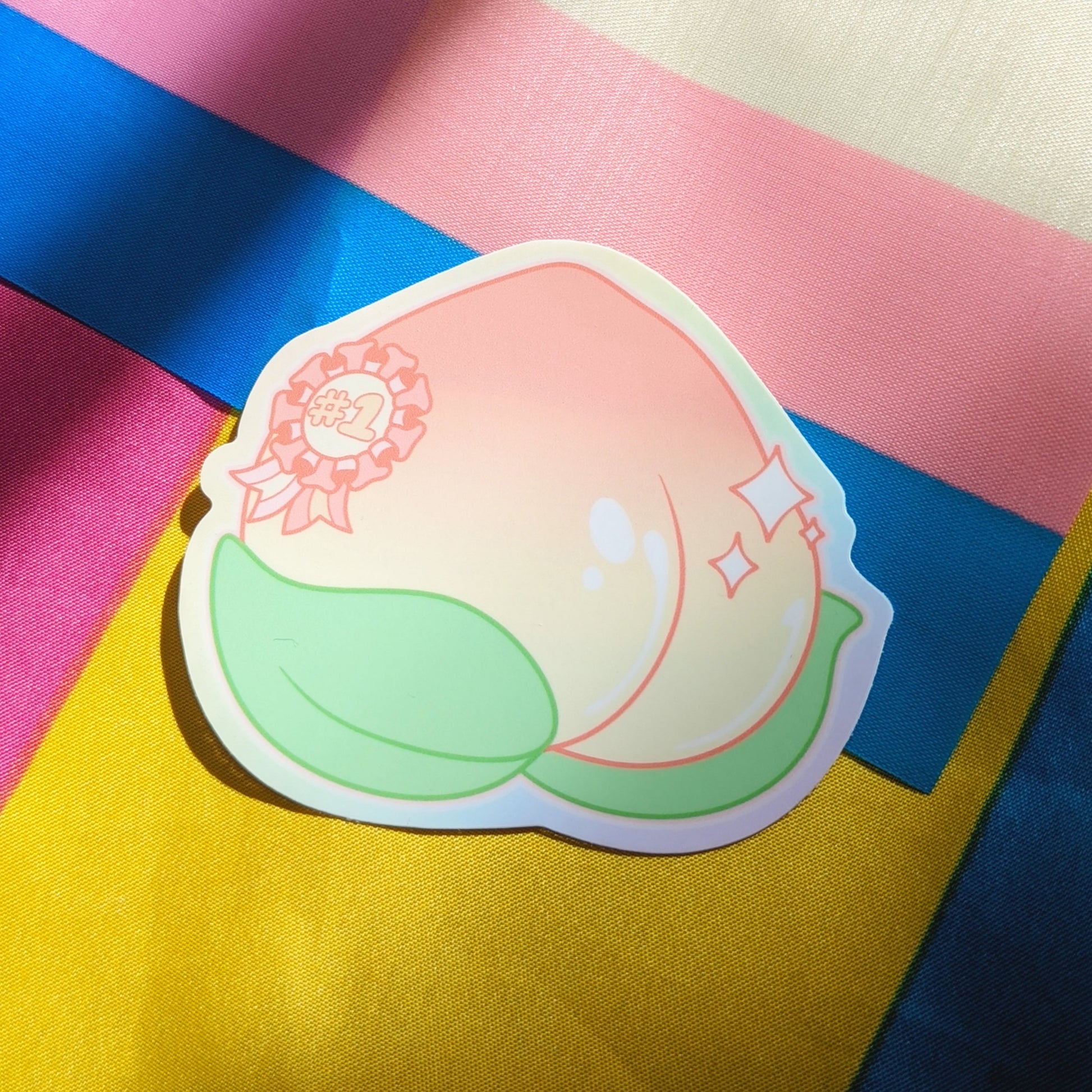 A photograph of a background with the transgender and pansexual pride flags. On top of the moss is a vinyl sticker of a shiny peach with a sparkly #1 ribbon on it.