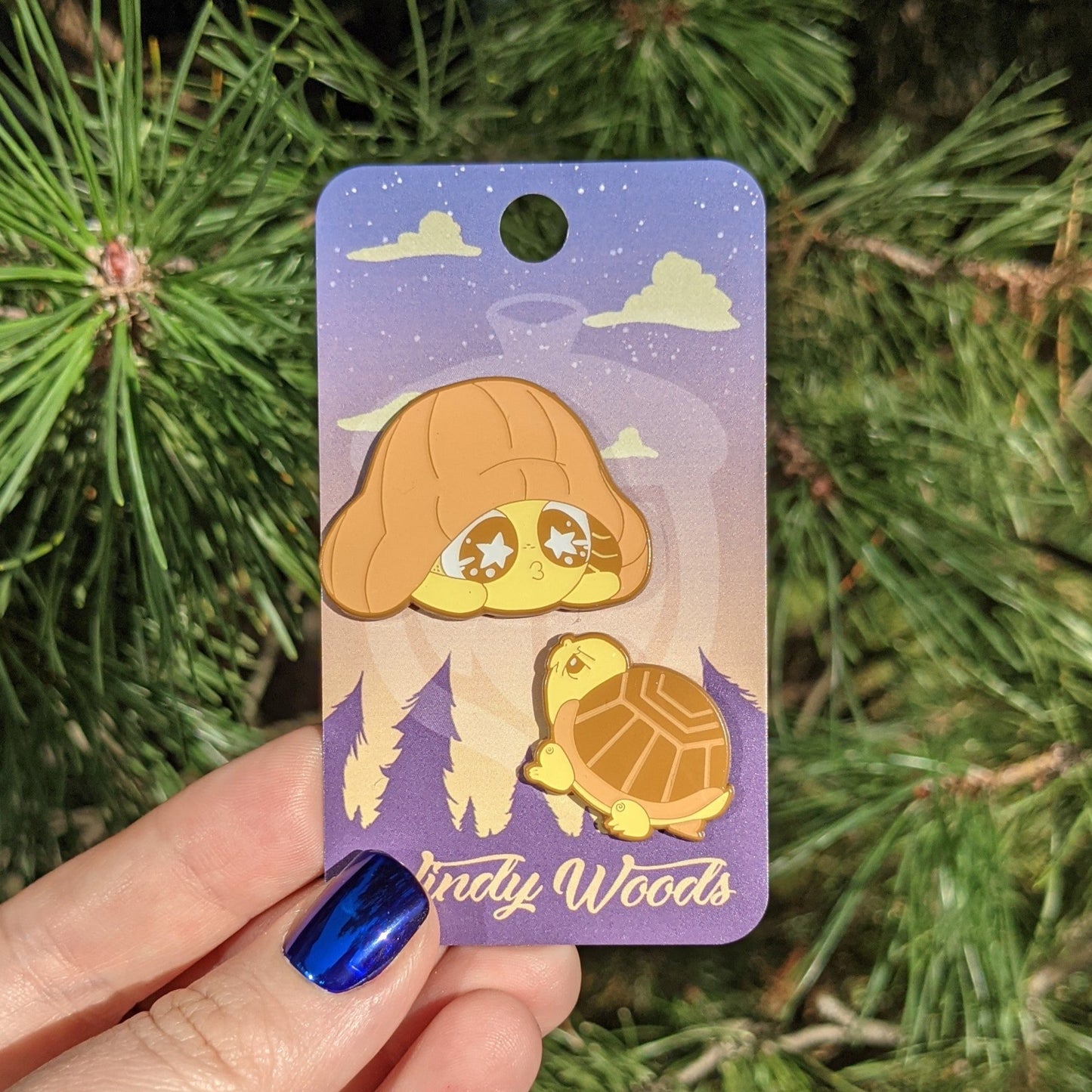 A photograph of an enamel pin being held in front of a pine tree. The enamel pin is on a purple Windy Woods backing card. The two pins on the card are an angry looking orange turtle looking at the second pin over an orange Mushkin mimicking the turtle with its cap.