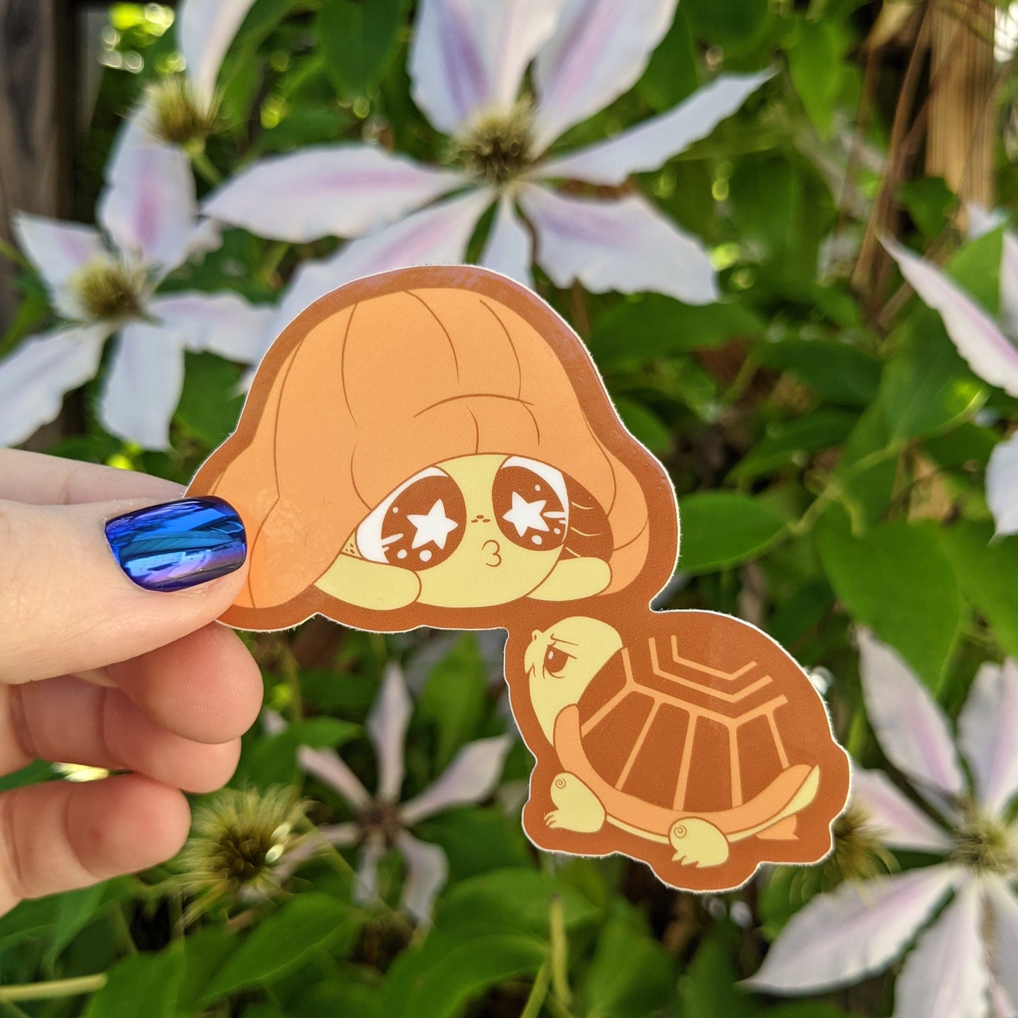 A photograph of a vinyl sticker being held in front of a clematis plant. The sticker is of an orange mushroom sprite called a Mushkin. Mushkin is mimicking a turtle by hiding under it’s mushroom cap.