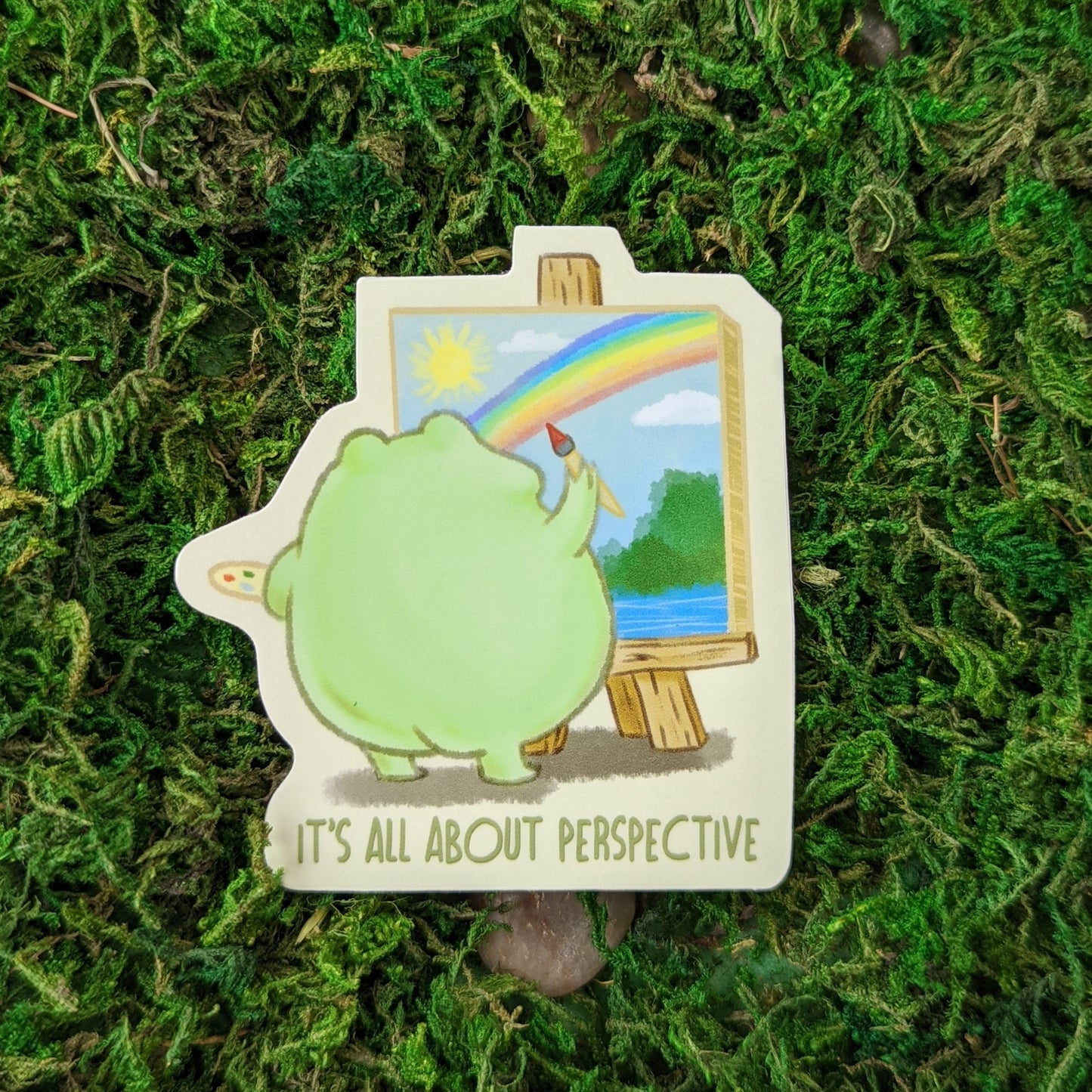 Photograph of a mossy background with a 3 inch vinyl sticker of a frog painting a picture of a rainbow. His little booty is adorable. Under the frog it says “It’s all about perspective.”