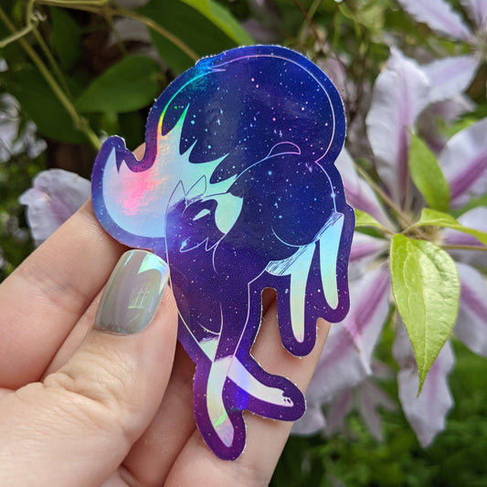 A photograph of a vinyl sticker being held in front of a clematis plant. The vinyl sticker is of a purple holographic galaxy themed fox.