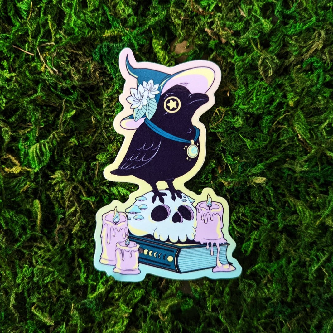 Photograph of a mossy background with a vinyl sticker of a raven witch with a teal and pink hat. The raven is standing on a skull on top of a book surrounded by pink candles.