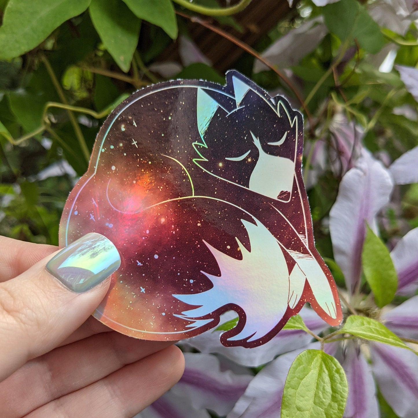 A photograph of a vinyl sticker being held in front of a clematis plant. The vinyl sticker is of a red holographic galaxy themed fox.
