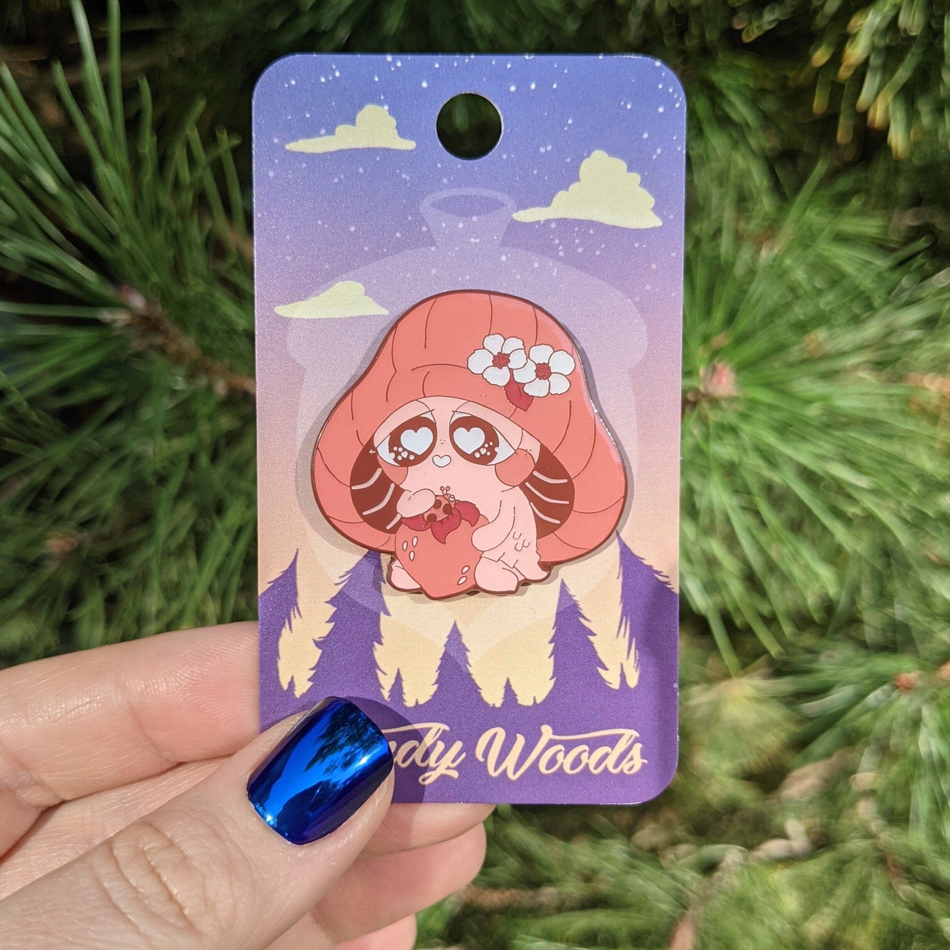 A photograph of an enamel pin on a backing card being held in front of some pine needles. The pin has artwork of a red mushroom sprite called a Mushkin. Mushkin is holding a strawberry with a ladybug on it.