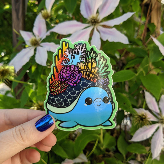 A photograph of a vinyl sticker being held in front of a clematis plant. The vinyl sticker of a blue turtle with coral growing out of its shell.