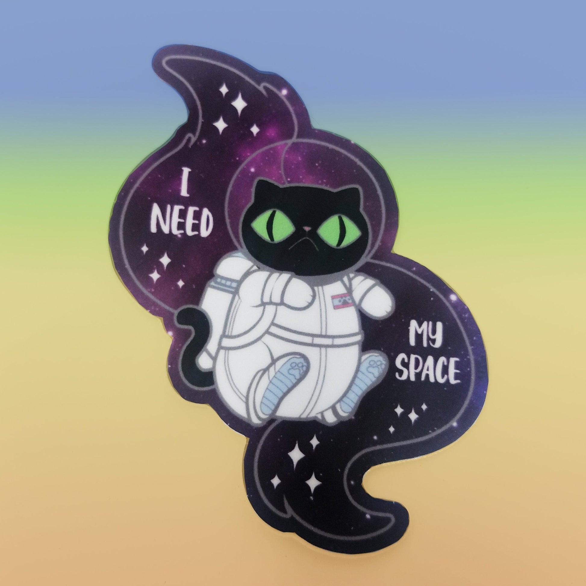 A photograph of a vinyl sticker resting on a pastel rainbow background. The vinyl sticker is of a black cat in a space suit floating through space. The sticker reads, “I need my space.”