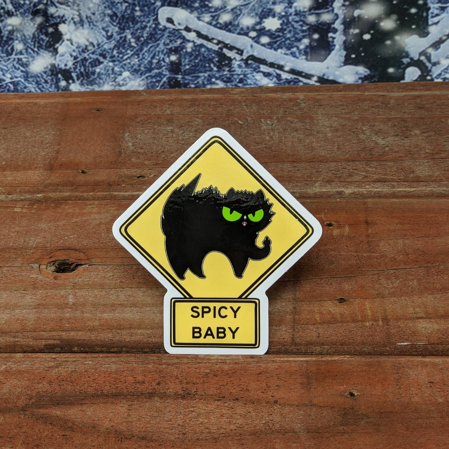 A redwood background with a snowy backdrop. The vinyl sticker is of a caution sign that reads, “Spicy Baby.” The sign has a poofed up black cat with green eyes. They are holding a front paw up and flipping the bird.