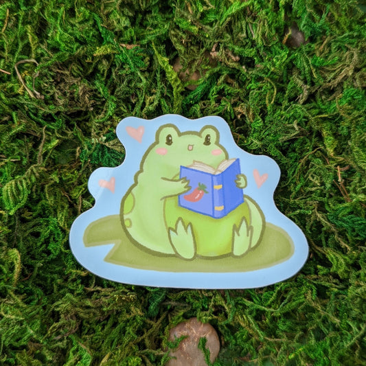 Photograph of a mossy background with a 3 inch vinyl sticker of a Frog on a lily pad holding a blue book with a spicy pepper indicating that they’re reading fan fiction.