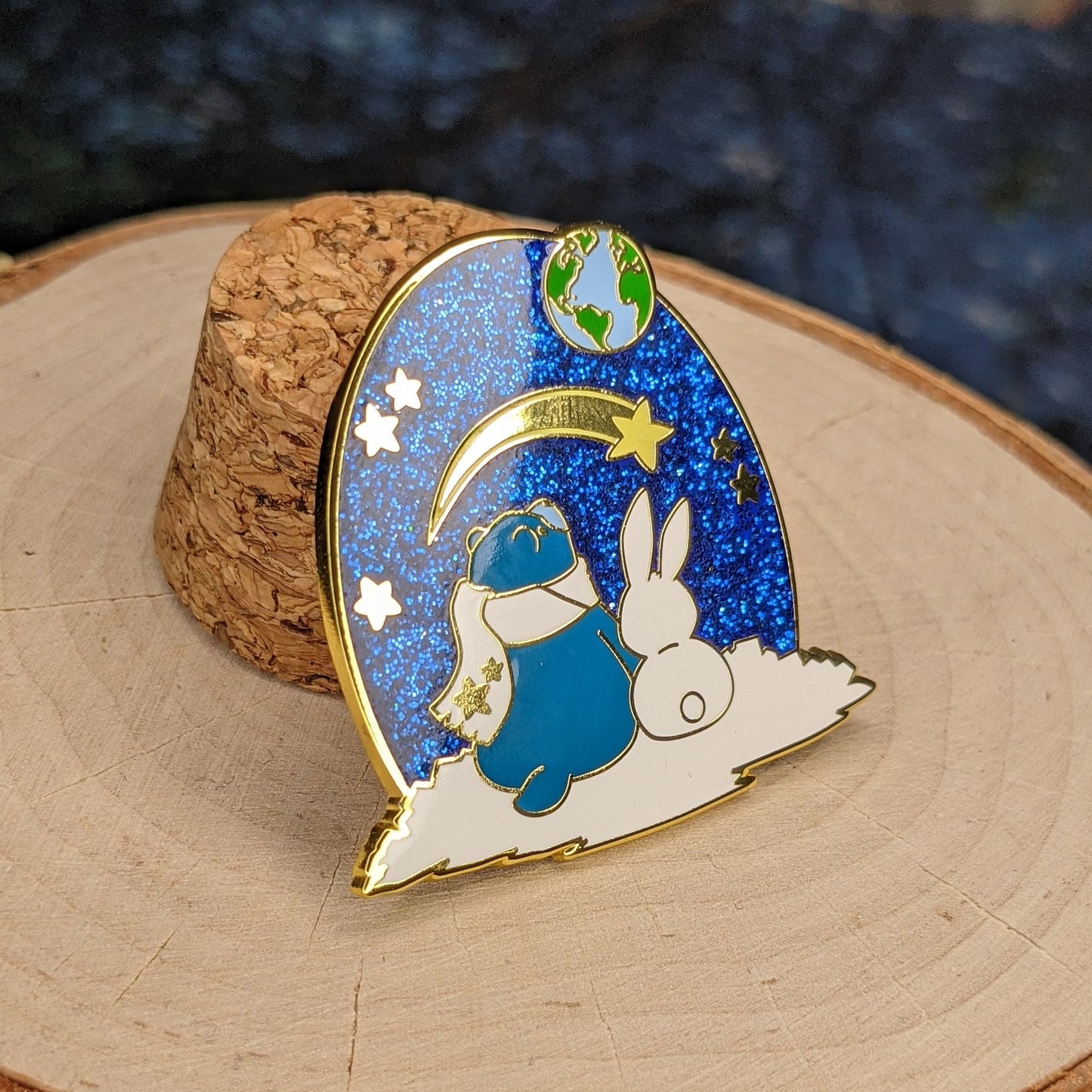 A photograph of a birch round and a cork holding up an enamel pin. The enamel pin artwork is of a blue star bear in a scarf and a white rabbit looking up at the Earth and a shooting star from the surface of the moon.