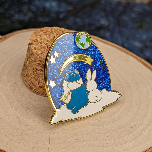 A photograph of a birch round and a cork holding up an enamel pin. The enamel pin artwork is of a blue star bear in a scarf and a white rabbit looking up at the Earth and a shooting star from the surface of the moon.