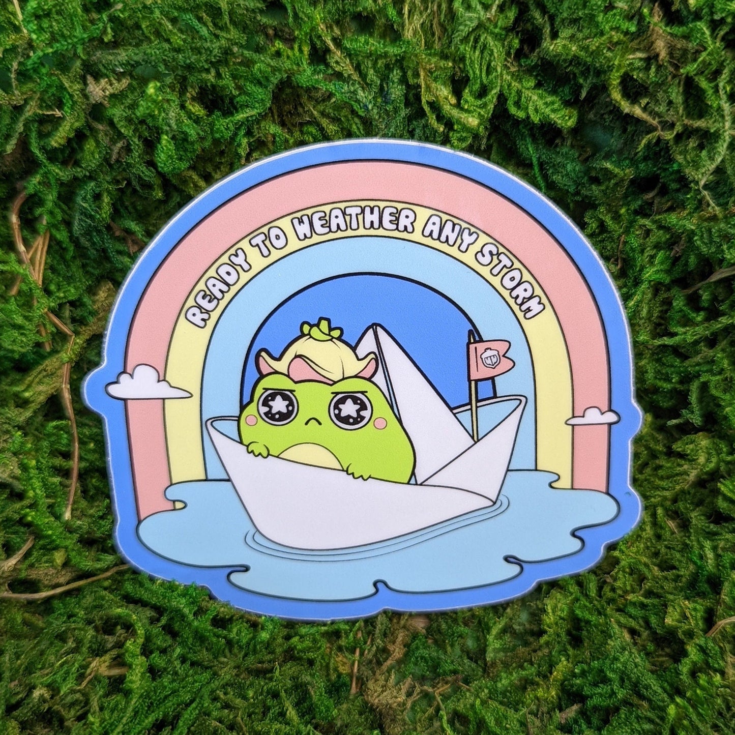 Photograph of a mossy background with a 3 inch vinyl sticker of a Frog in a paper boat floating under a rainbow. The text says “Ready to weather any storm.”
