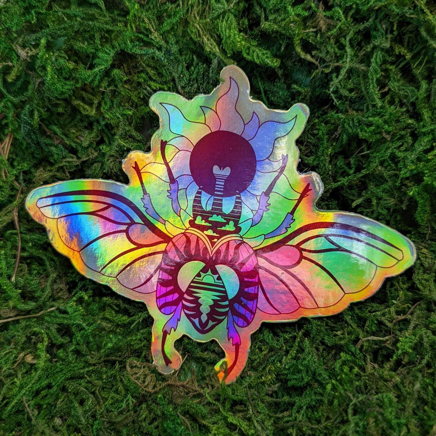 Photograph of a mossy background with a vinyl sticker of a holographic beetle with a sun theme.