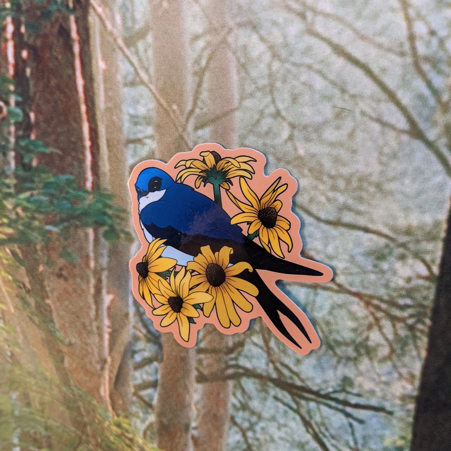 A photograph of a sticker being held in front of a forest tapestry. The sticker features a blue swallow bird hanging out with some black eyed susan flowers.