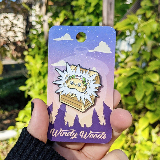 A photograph of an enamel pin being held in front of a clematis plant. The enamel pin is a treasure chest with a smiley faced taco coming out of it.