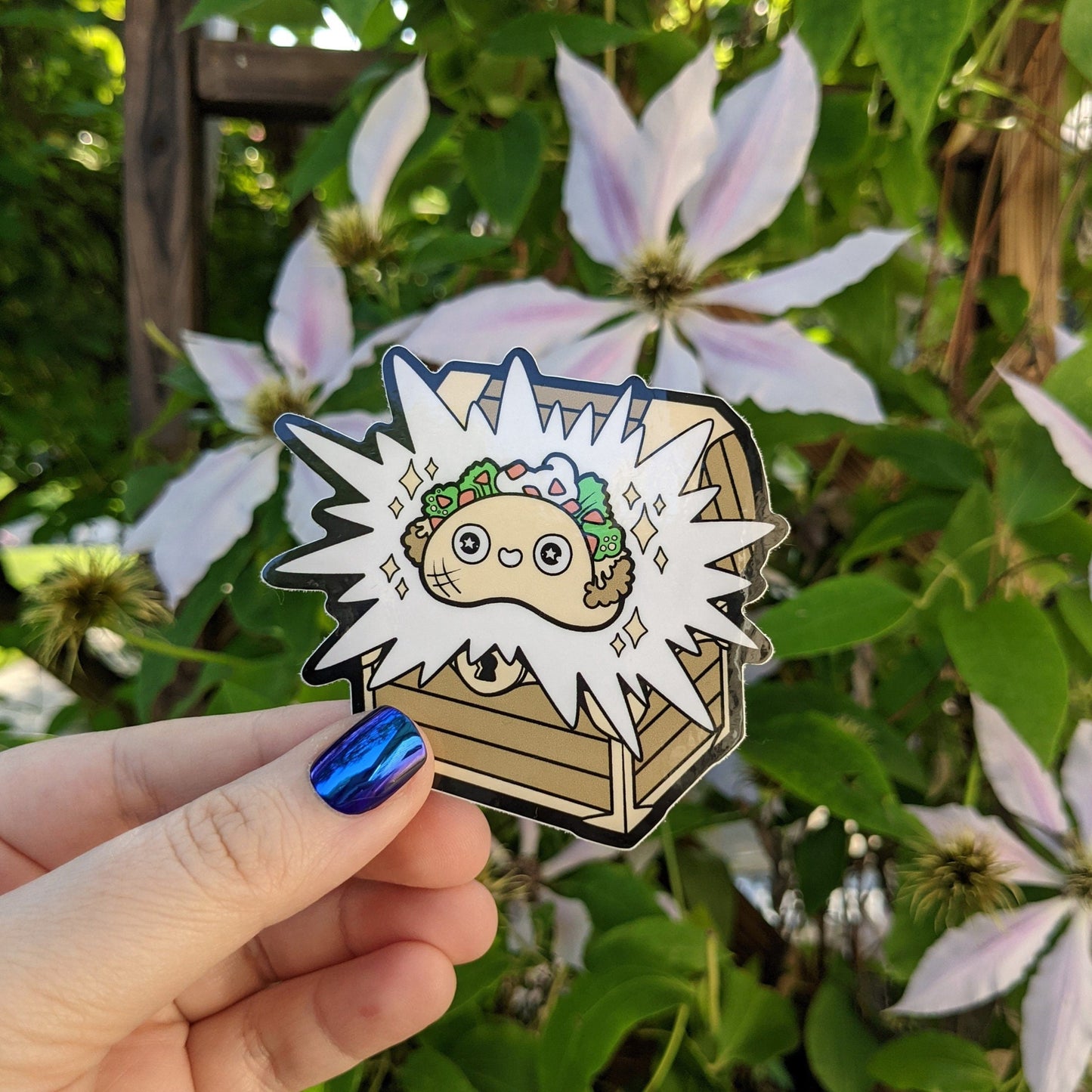 A photograph of a vinyl sticker being held in front of a clematis plant. The vinyl sticker is a treasure chest with a smiley faced taco coming out of it.