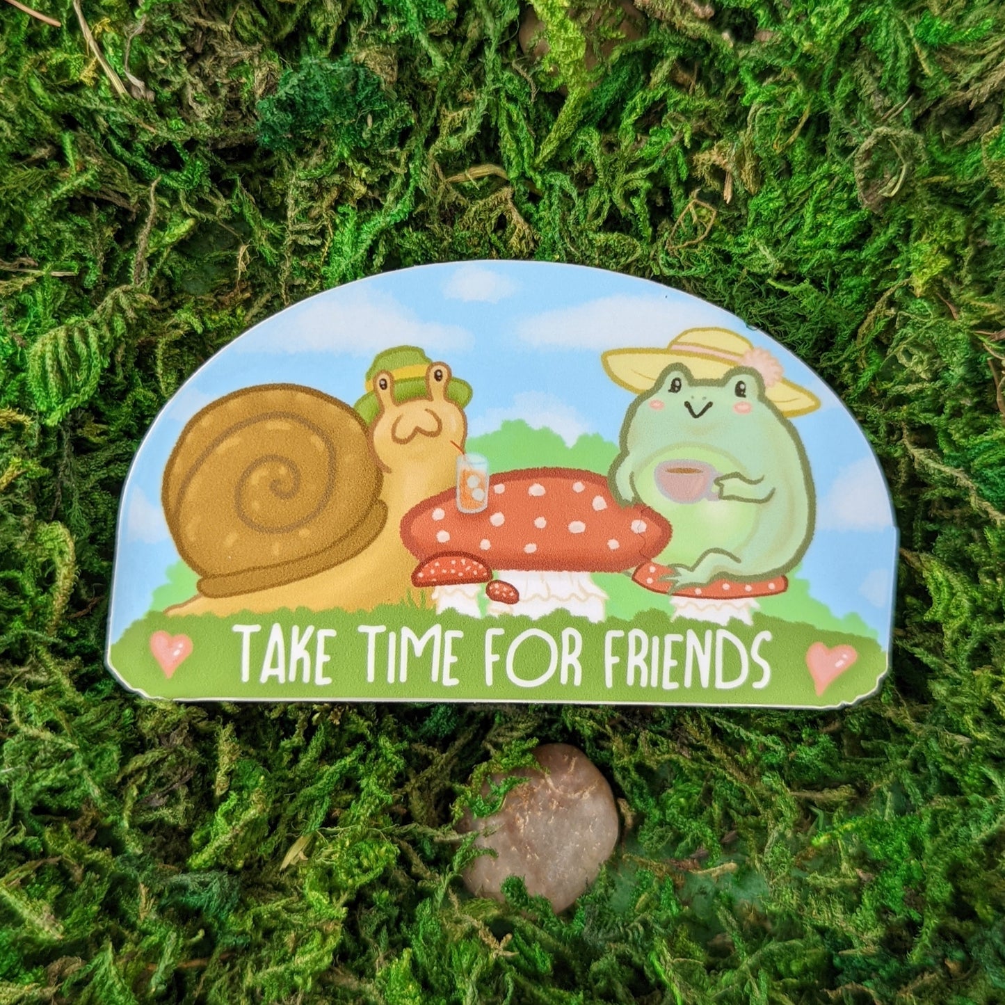 Photograph of a mossy background with a 3.5 inch vinyl sticker of a Frog and a snail having tea and coffee on a mushroom table. 