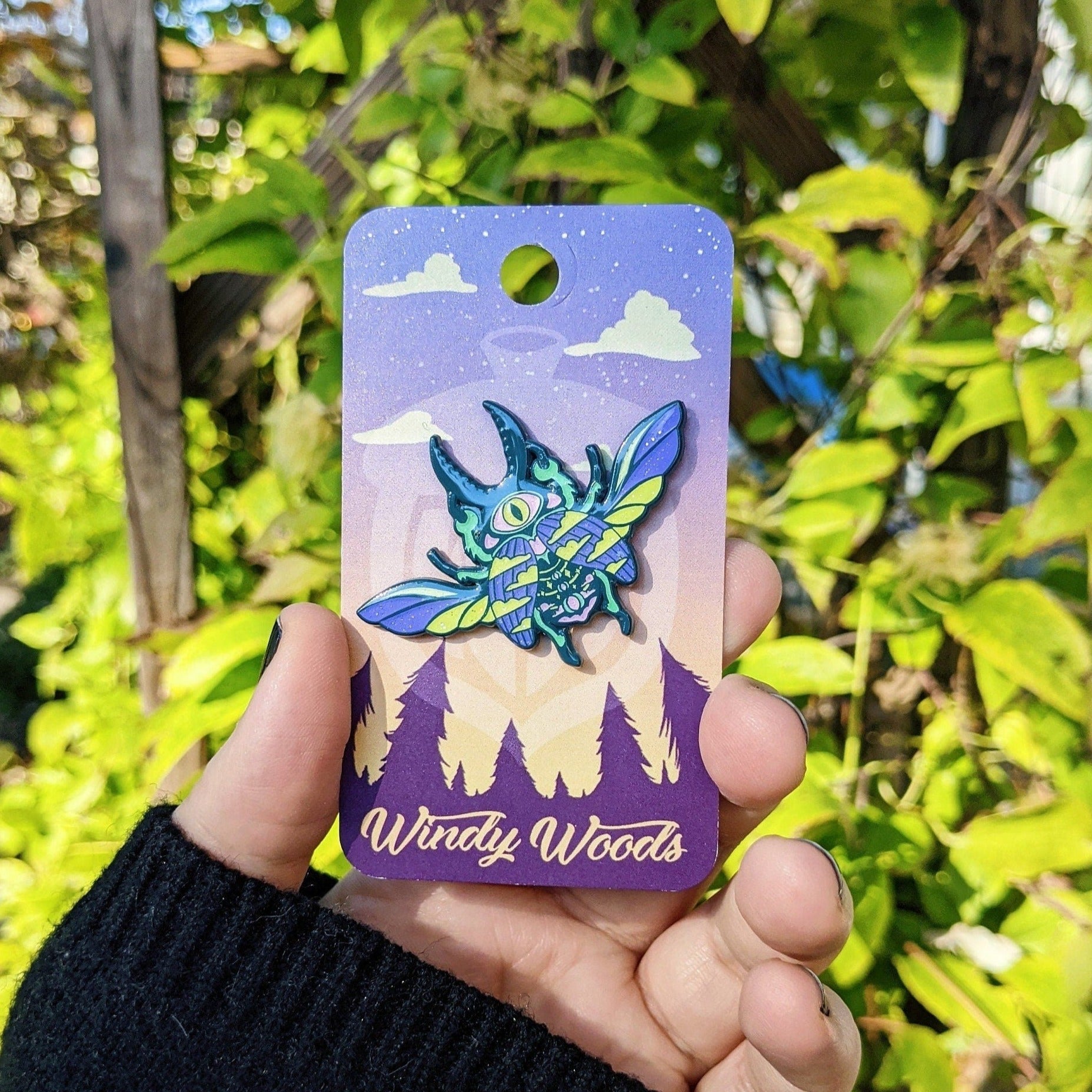 A photograph of an enamel pin being held in front of a clematis plant. The enamel pin features a scarab beetle with one eye. The colors scheme of the beetle is purple, chartreuse, and teal.